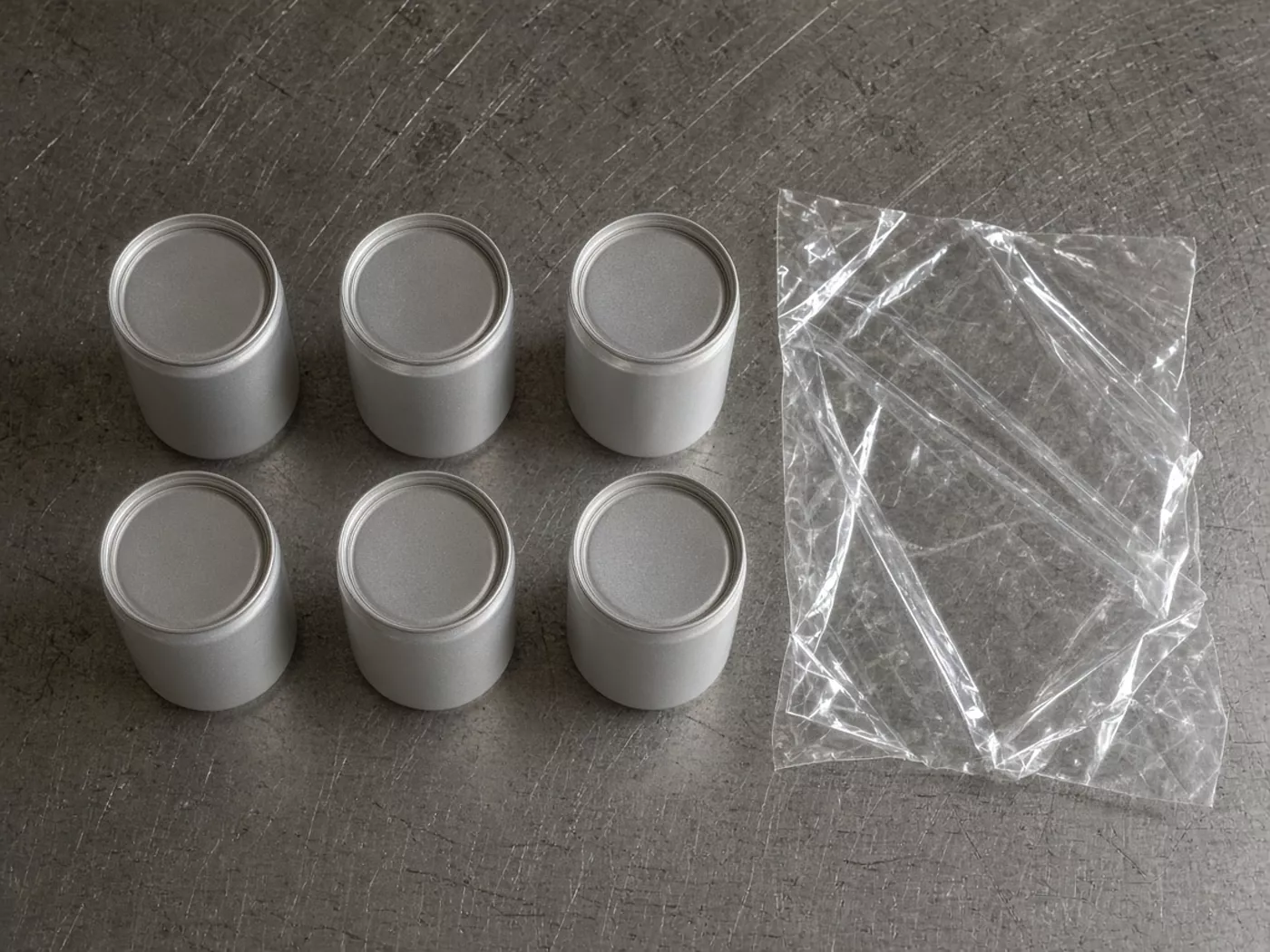 Six plain cans in two rows on a scratched steel table, beside a loose sheet of clear film